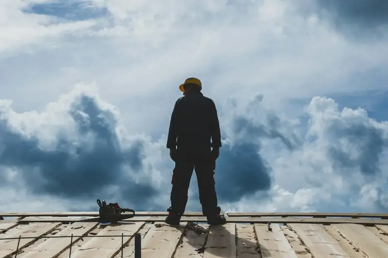Couvreur vu de dos, debout sur un toit en chantier face à un grand ciel chargé de nuages
